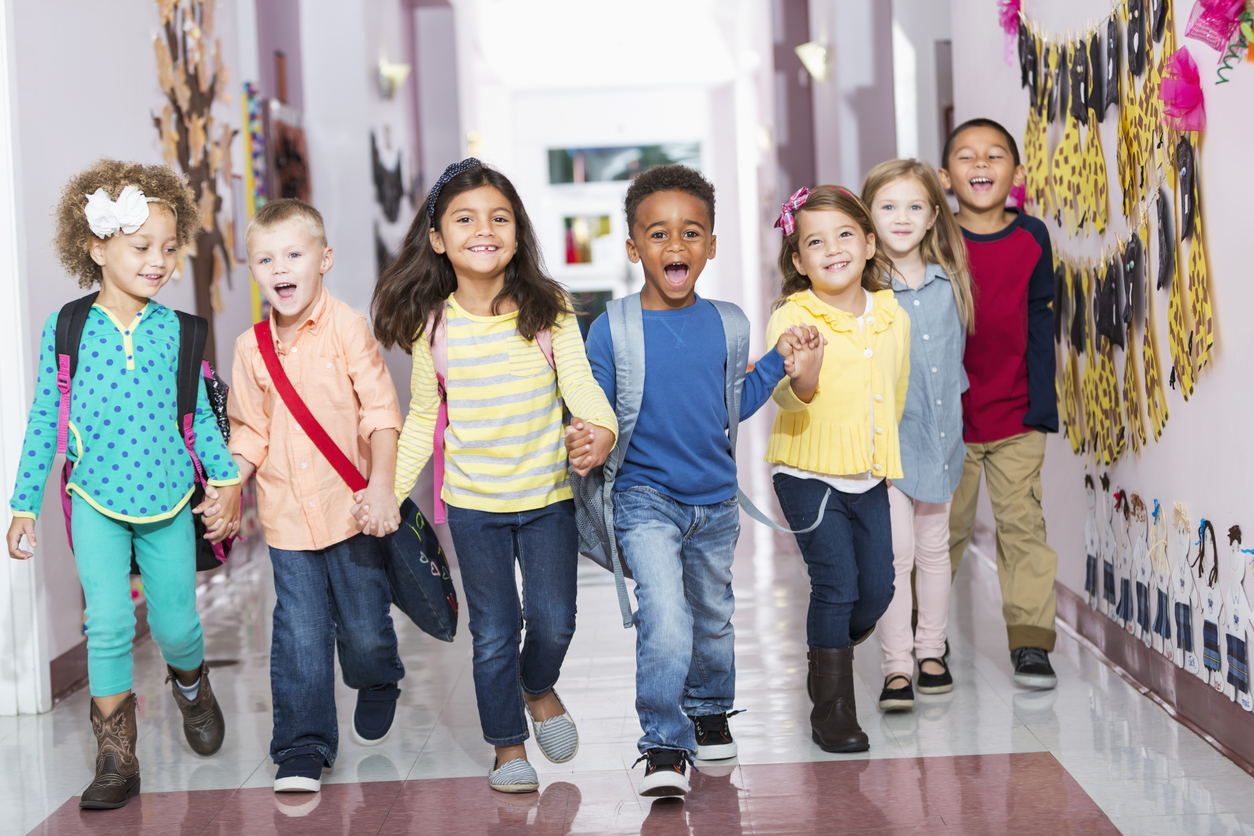 七个多民族的孩子手牵着手, 跑过学校的走廊, 笑着喊着, 看着镜头. 小男孩和女孩是幼儿园或学前班的年龄，4到6岁.