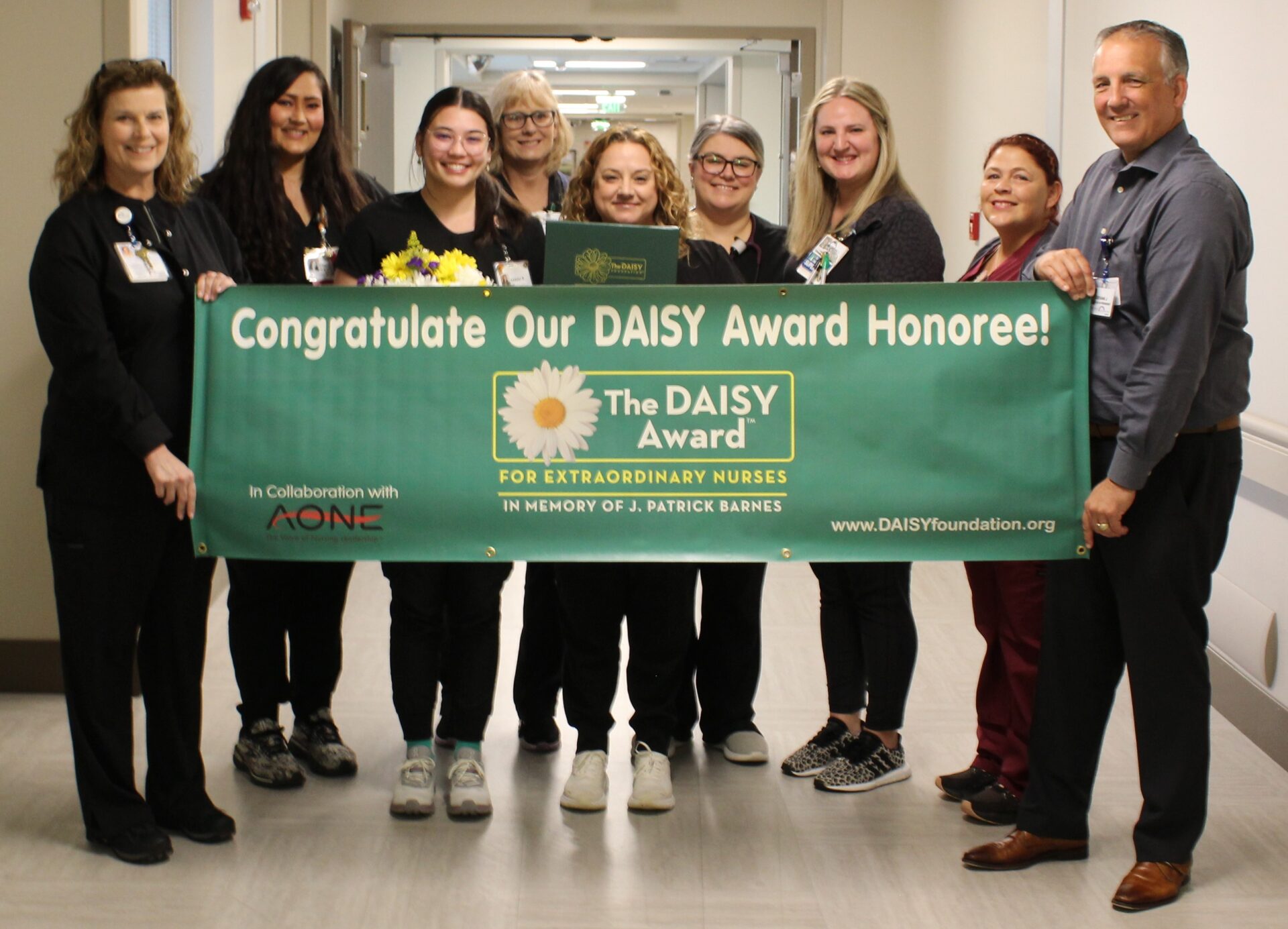 Sarah pictured with her co-workers and leadership holding green DAISY banner.