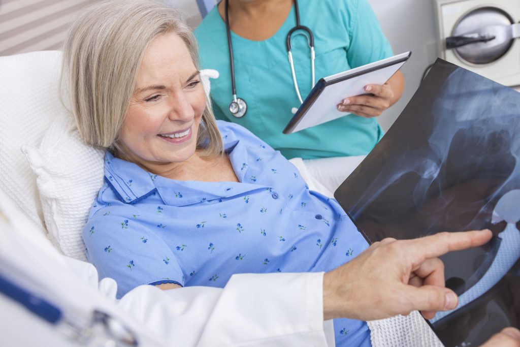 Happy senior woman patient recovering in hospital bed with male doctor and female nurse looking at hip replacement x-ray