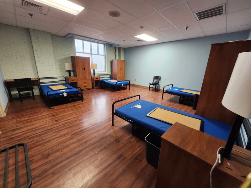 A dorm-style room with four blue beds, four wooden wardrobes, desks, black chairs, a lamp, and a window. The floor is wood, and the walls are light blue and brick. The room is clean, simple, and spacious.