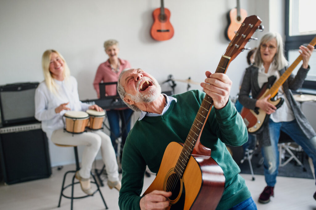 A joyful elderly man plays acoustic guitar energetically in front of three smiling older women—one playing bongo drums, one with an electric guitar, and one standing happily—in a bright room decorated with guitars. Their lively music and cheerful camaraderie highlight how engaging group activities can positively impact individuals living with Alzheimer’s, emphasizing the difference between Alzheimer’s disease and dementia through moments of shared joy and connection.