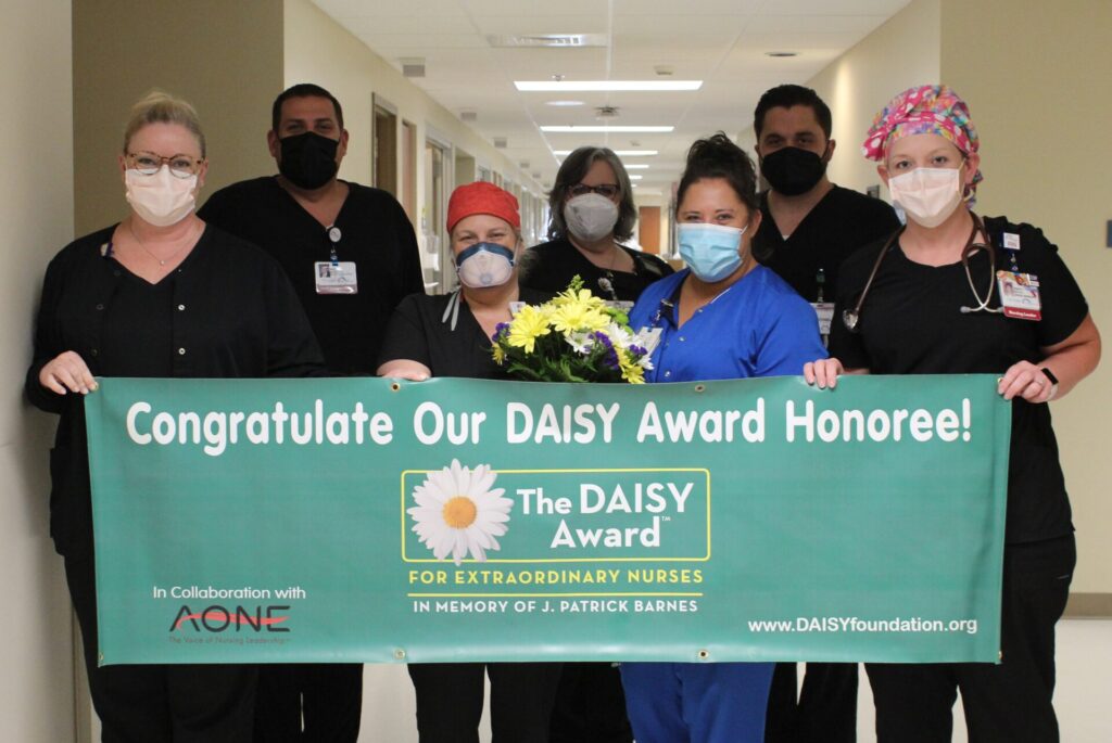 Seven healthcare workers in scrubs and masks stand in a hospital hallway, holding a green banner that says, Congratulate Our DAISY Award Honoree! The DAISY Award for Extraordinary Nurses.