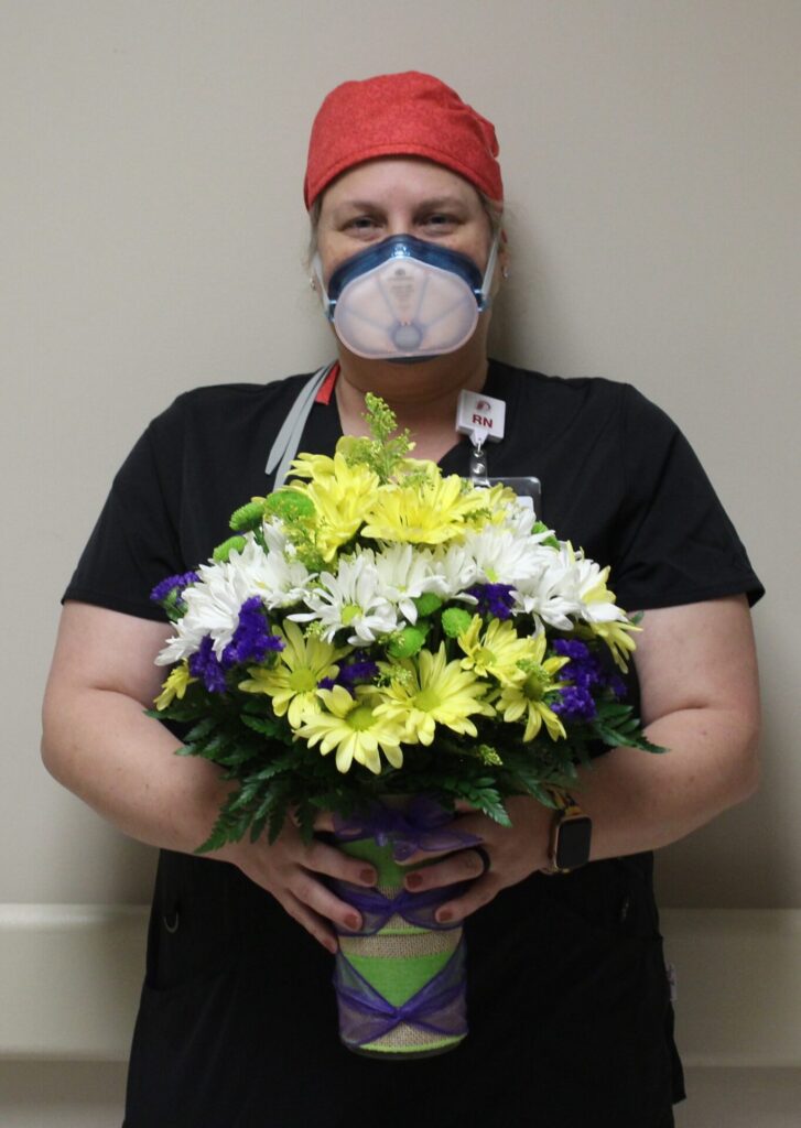 A nurse in black scrubs and a red cap, wearing a clear face mask, stands against a beige wall while holding a large bouquet of yellow, white, and purple flowers with green leaves.