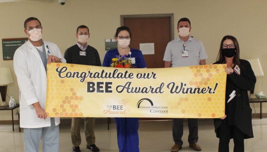 Five healthcare workers, all wearing masks, stand indoors holding a yellow banner that reads Congratulations our BEE Award Winner! One person in the center holds a bouquet of flowers.