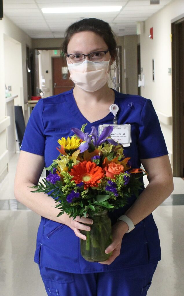 A nurse in blue scrubs, wearing glasses and a face mask, stands in a hospital hallway holding a colorful bouquet of flowers in a glass vase.