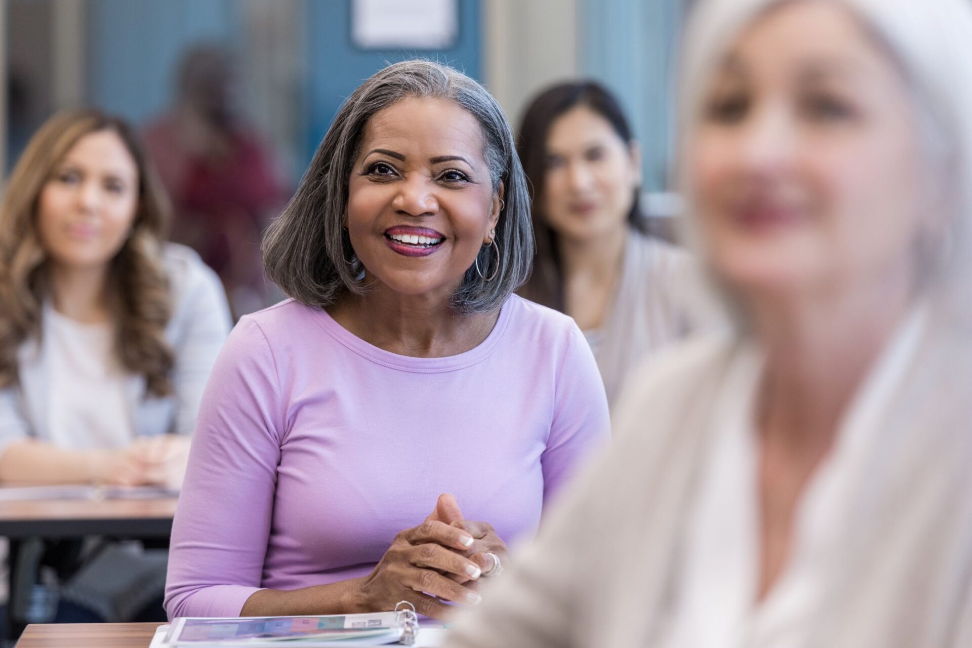 A smiling older woman with gray hair sits at a desk in a classroom at Parkwest Medical Center, surrounded by other women of varying ages, all appearing attentive and engaged.