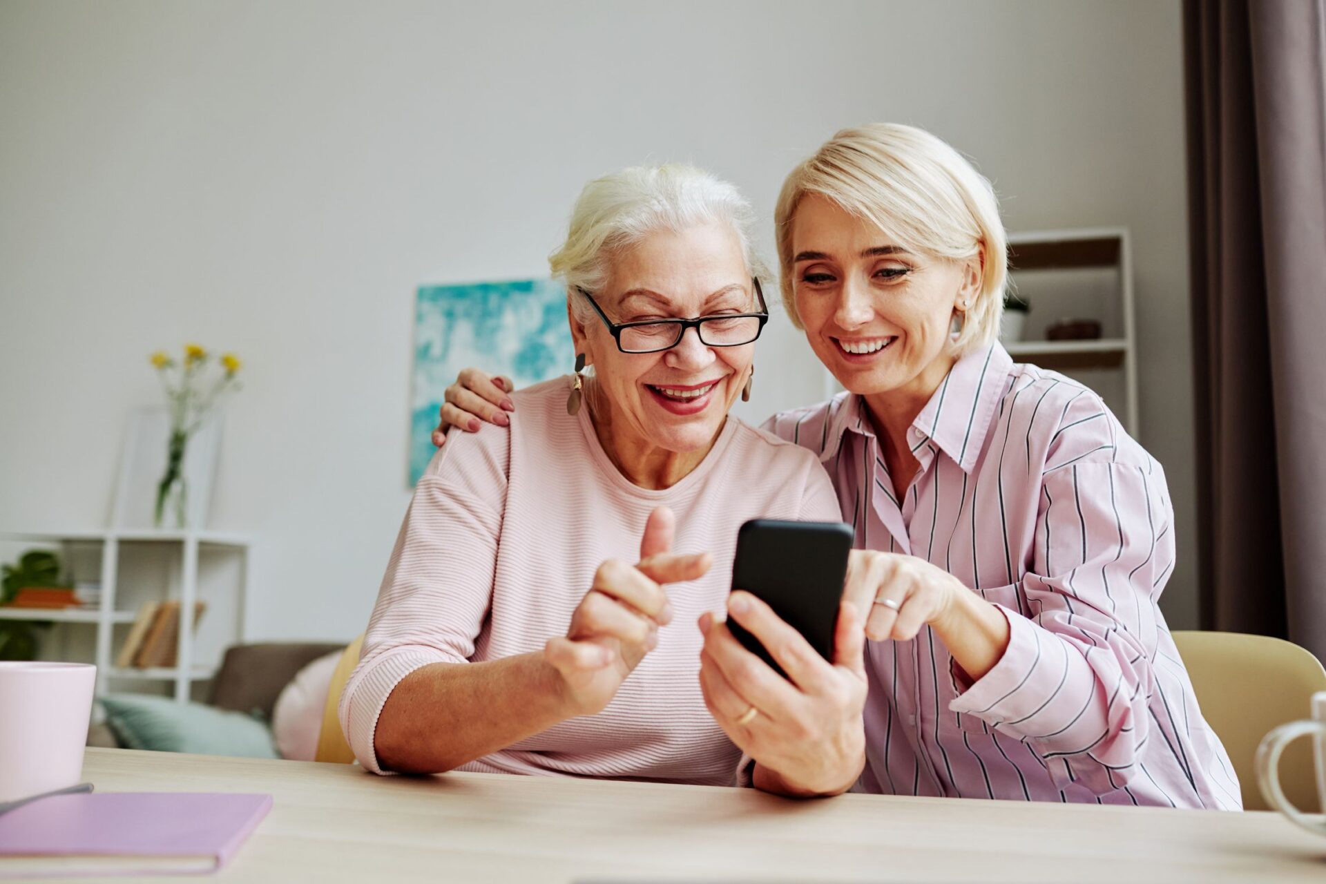 Two women, one older and one younger, sit together smiling and looking at a smartphone. The younger woman is pointing at the phone, while the older woman, wearing glasses, enjoys the moment. They appear happy and engaged at Parkwest Medical Center.