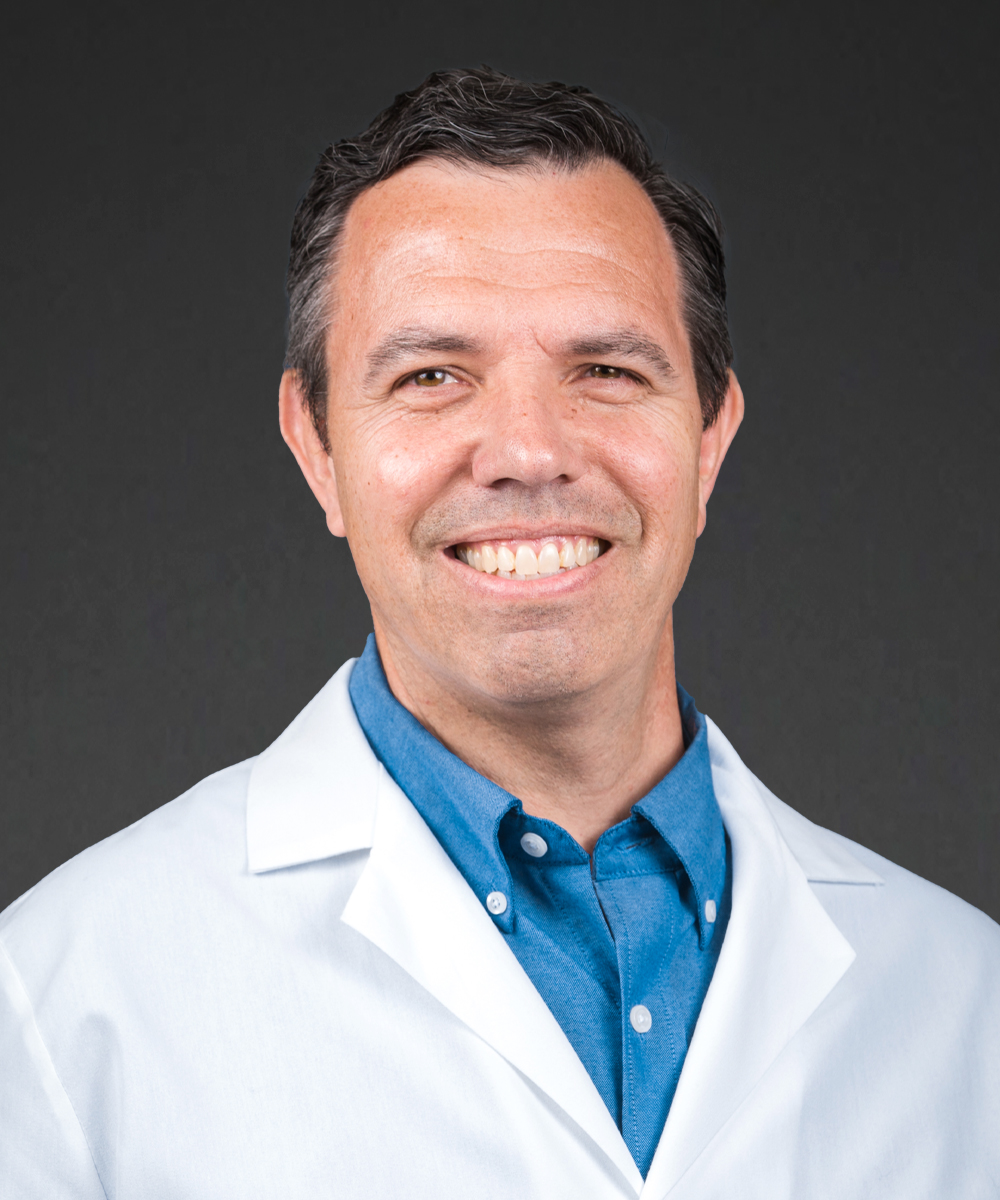 A man wearing a white lab coat and blue shirt smiles at the camera against a plain dark background, embodying the expertise and professionalism of medcare specialists.