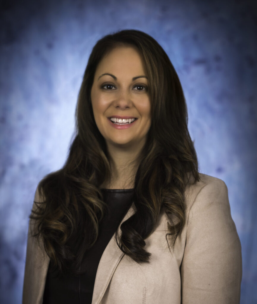 A woman with long brown hair wearing a beige blazer over a black top is smiling at the camera, set against a blue, softly blurred backdrop, capturing warmth and approachability in just about every detail.