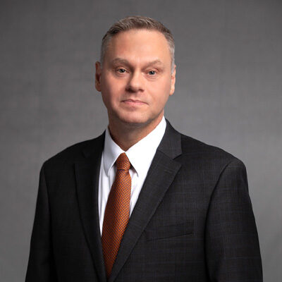 A man in a dark suit, white dress shirt, and orange patterned tie stands about center in front of a plain gray background, facing the camera with a neutral expression.