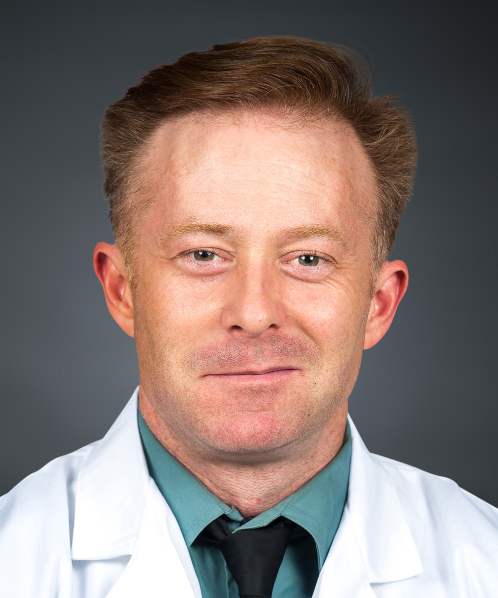 A man with short light brown hair wearing a white lab coat, teal shirt, and black tie, smiling slightly in front of a dark grey background, embodying the professionalism of medcare specialists.