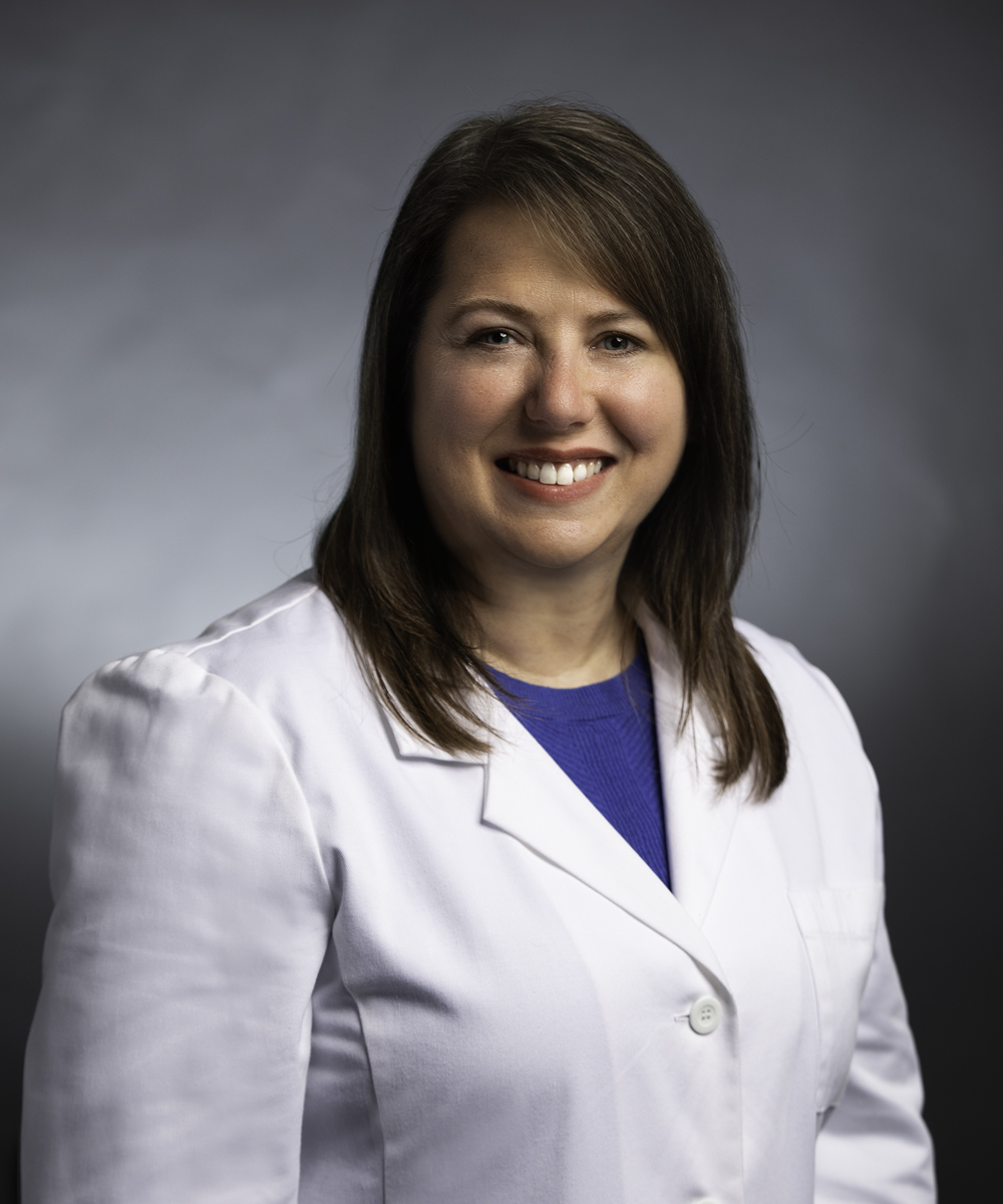 A smiling woman with straight brown hair, one of the dedicated medcare specialists, wears a white lab coat over a blue top, standing against a gray background.