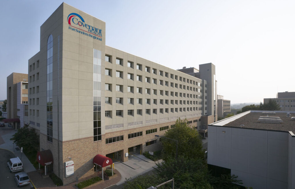 A large multi-story hospital building with a tan facade, displaying the sign Covenant Fort Sanders Hospital. About the area, trees and smaller buildings are nearby, and vehicles are parked along the surrounding streets.