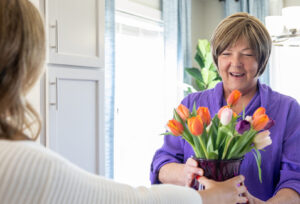 An older woman smiles as she receives a vase of colorful tulips from another person indoors. Sunlight streams through the window, brightening the cheerful scene.