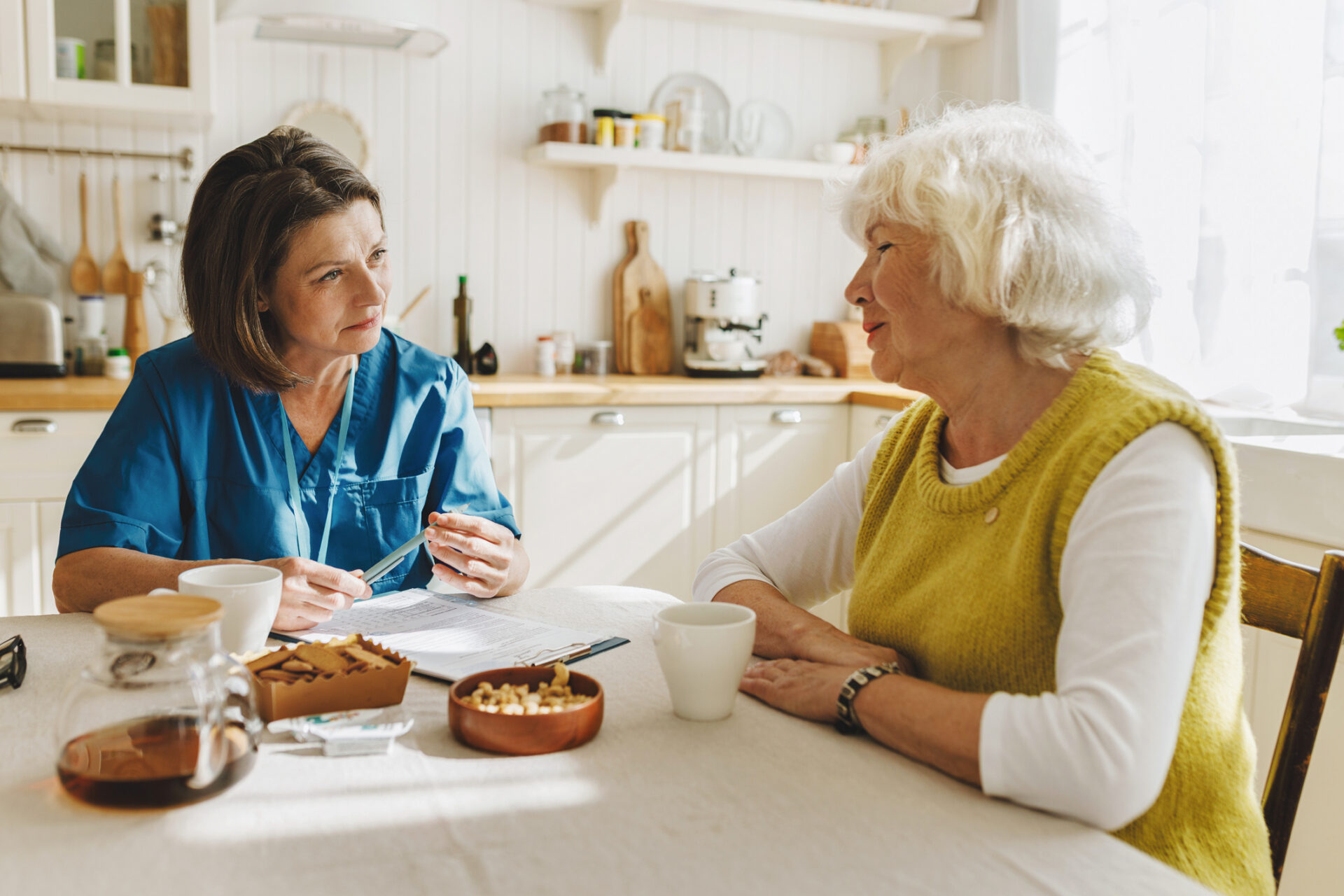 A nurse in blue scrubs sits at a kitchen table, attentively talking with an elderly woman in a yellow sweater. Papers, snacks, and coffee cups are on the table in a warmly lit kitchen.