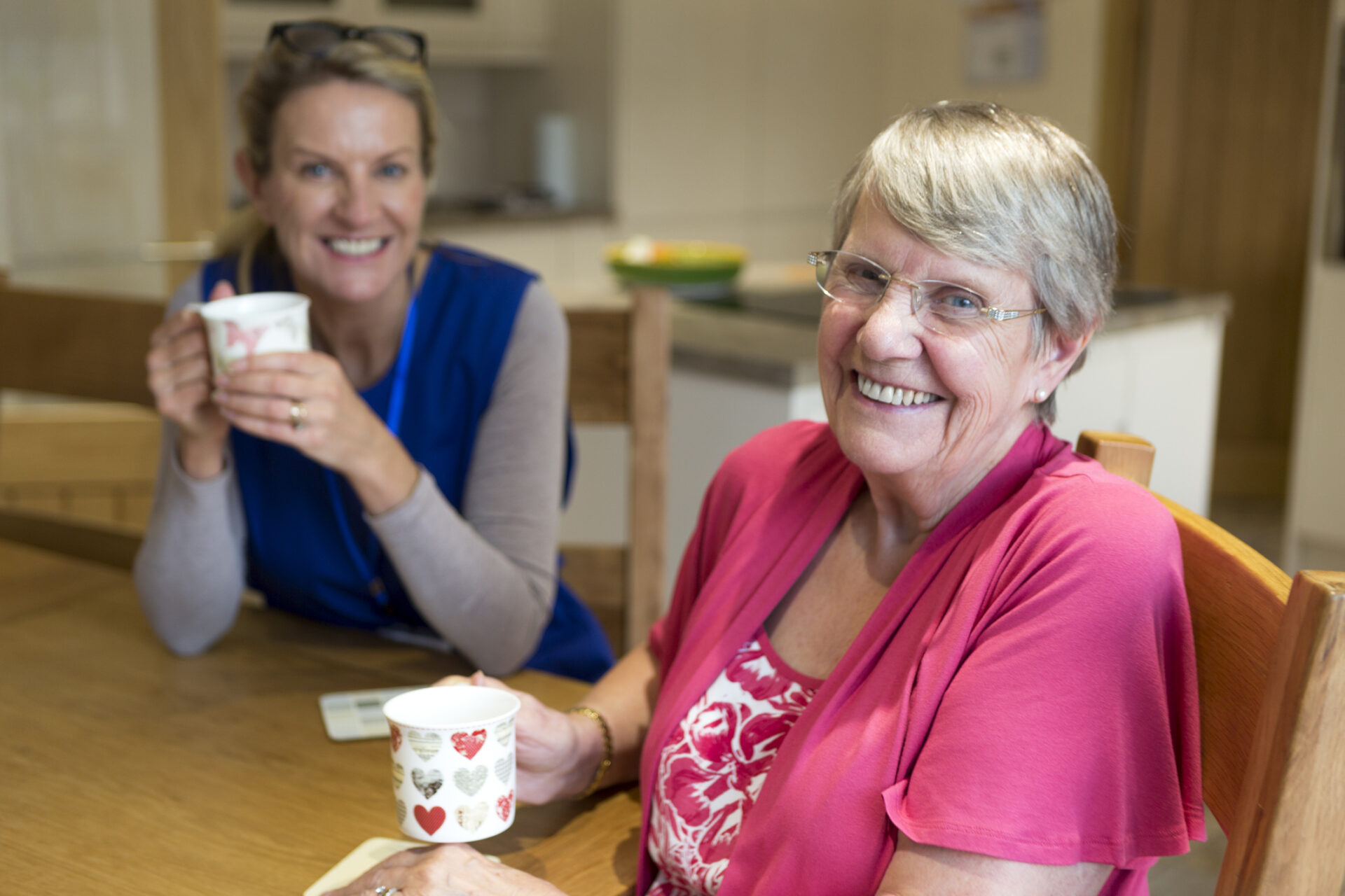 Two women sitting at a table, smiling and holding mugs. One woman wears glasses and a pink top, the other wears a blue vest. They appear to be enjoying a friendly conversation in a bright kitchen.