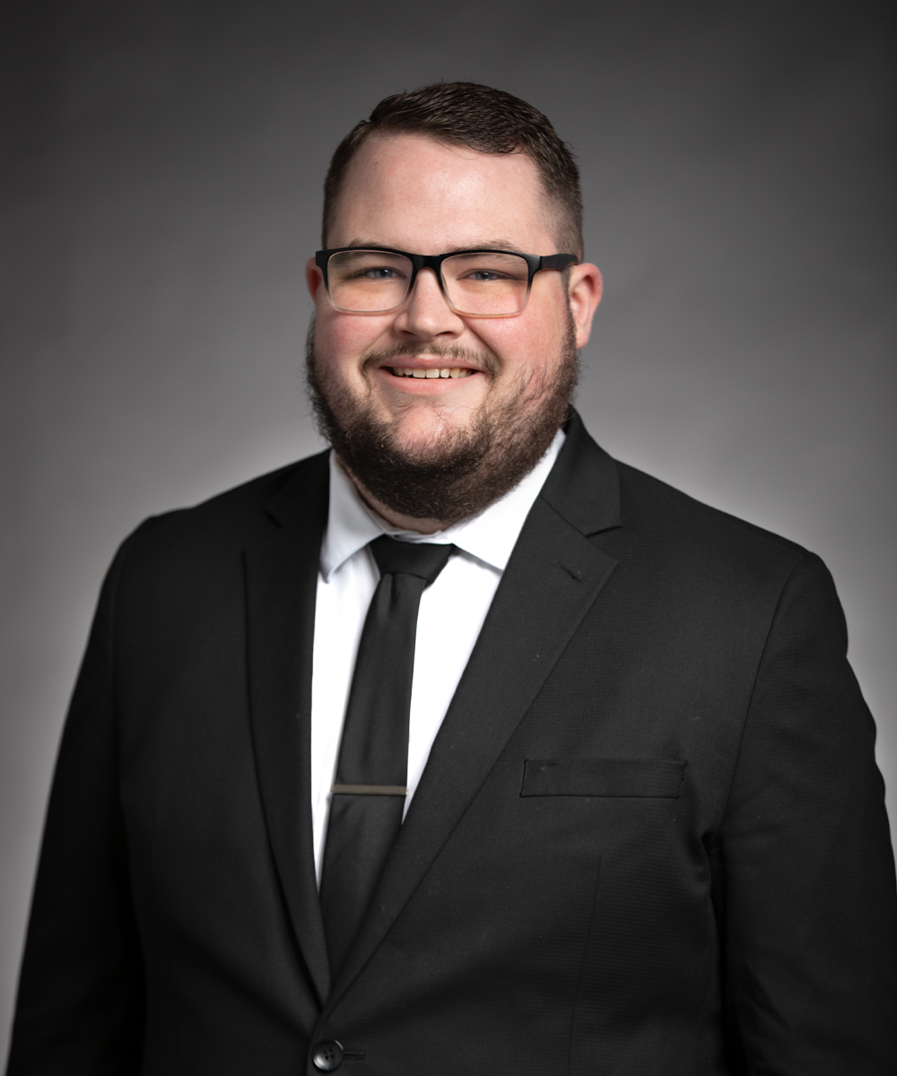 A man with short brown hair, glasses, and a beard, wearing a black suit, white shirt, and black tie, smiles at the camera against a plain gray background—perfect for promoting nursing jobs at Parkwest Medical Center.