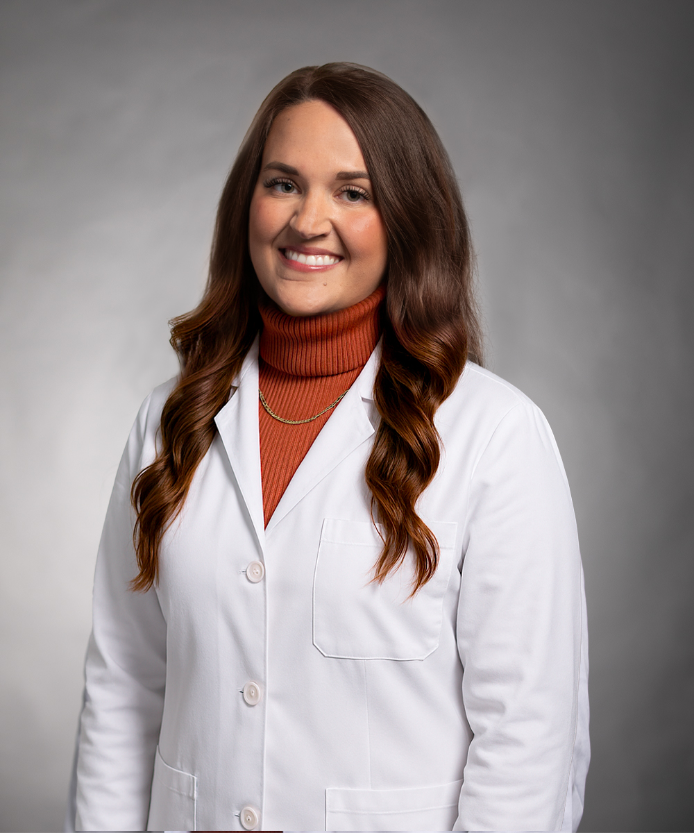 A woman with long brown hair in loose waves, wearing a white lab coat over an orange turtleneck sweater and a gold necklace, smiles while standing against a neutral gray background, representing women's care in Morristown.