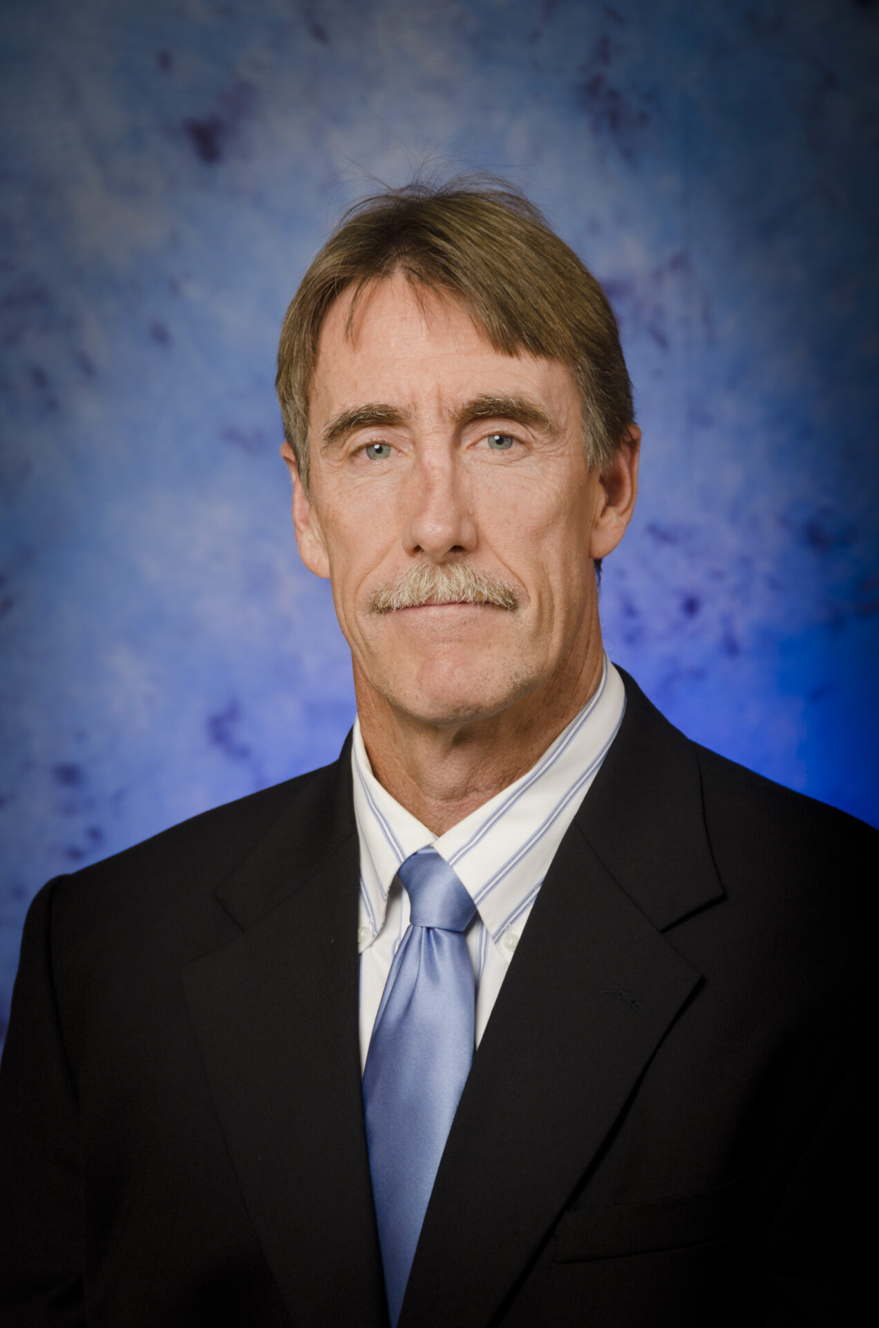 A middle-aged man with light brown hair and a mustache, dressed in a black suit, white shirt, and light blue tie, poses in front of a blue mottled backdrop—an advocate for womens care in Morristown.