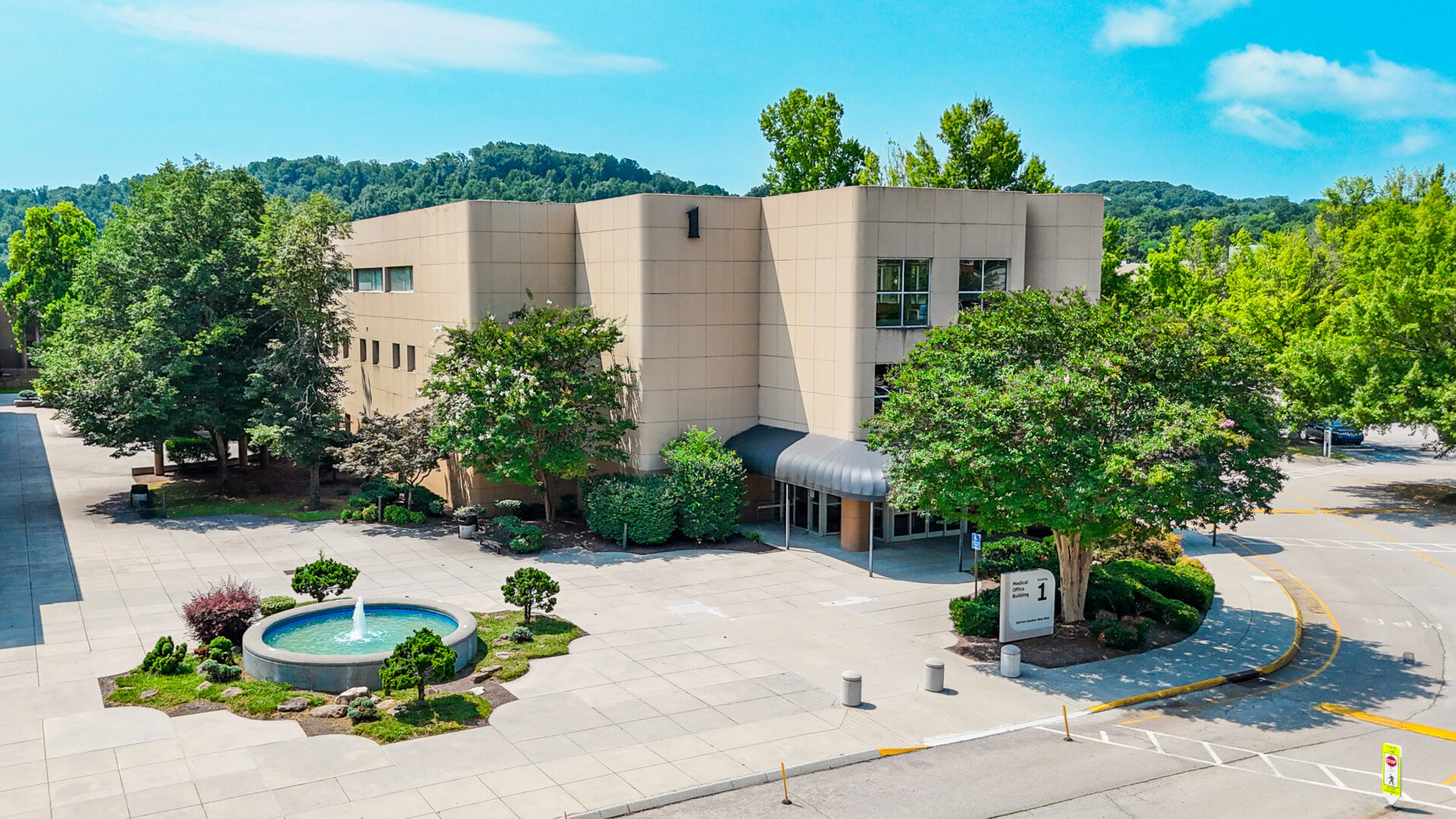 Modern beige building with large windows, surrounded by trees, serves as a welcoming therapy center in West Knoxville. A small fountain and landscaped area are in front, with a driveway and parking spaces nearby. Blue sky and green hills are visible in the background.