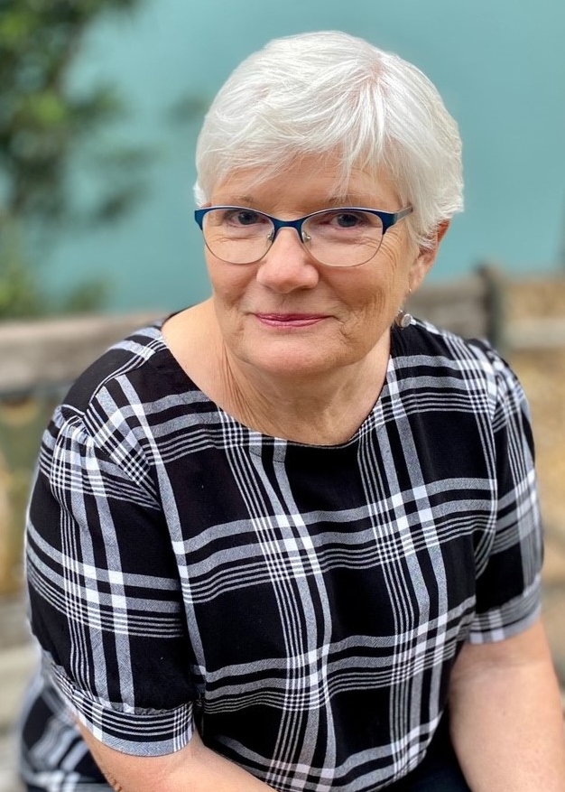 Older woman with short white hair and glasses, wearing a black plaid shirt, sits outdoors on a bench with a soft, green background at the Survivors of Suicide Loss Day Event.
