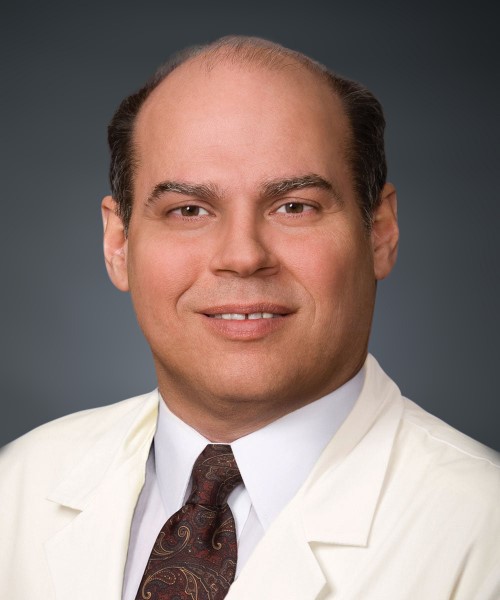 A man with light skin, short dark hair, and a receding hairline, wearing a white medical coat and patterned tie, poses in front of a plain dark background.