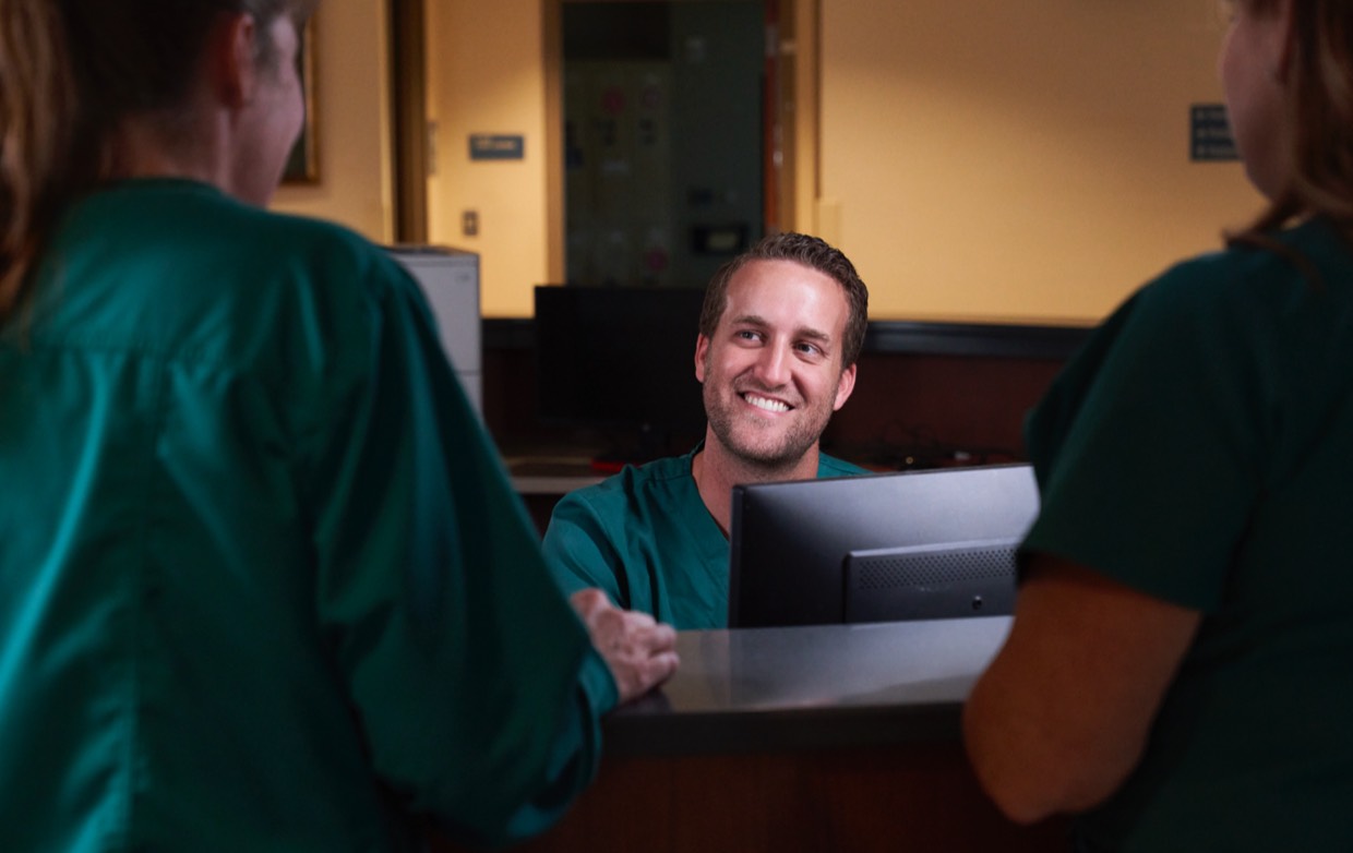 male nurse in scrubs smiling at computer while talking to two people