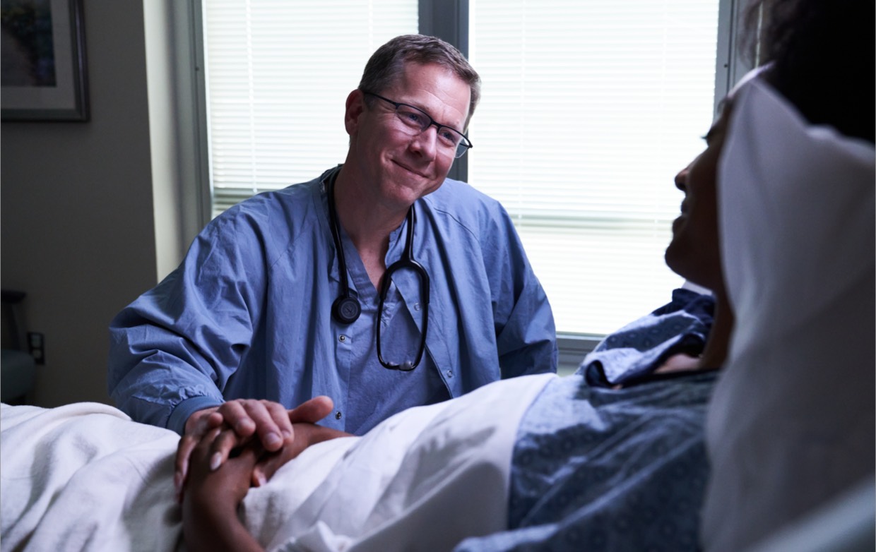 A middle age male doctor comforting a female patient who's in a hospital bed.