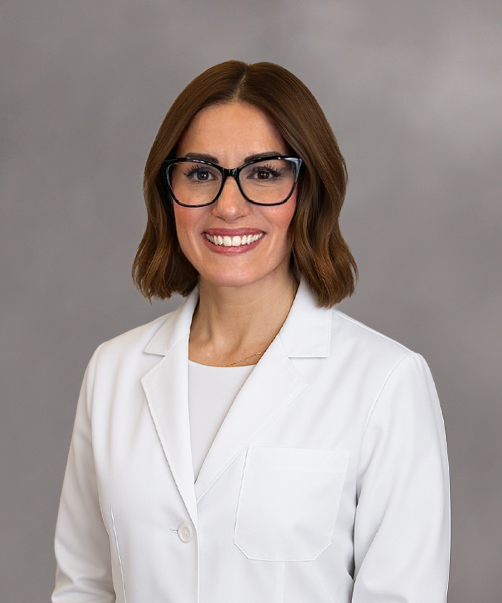 A woman with shoulder-length brown hair and glasses smiles, wearing a white lab coat—representing women's care in Morristown—against a plain gray background.