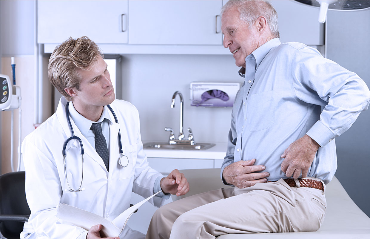 young male doctor listening to senior male patient sitting on bed indicating hip