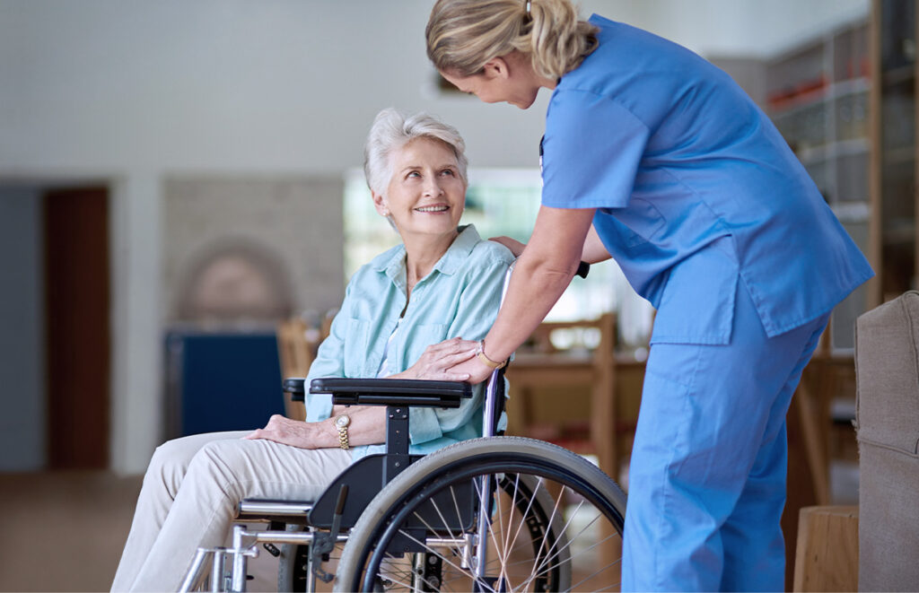 A nurse in blue scrubs smiles and gently holds the hand of an older woman sitting in a wheelchair inside a bright, comfortable room.