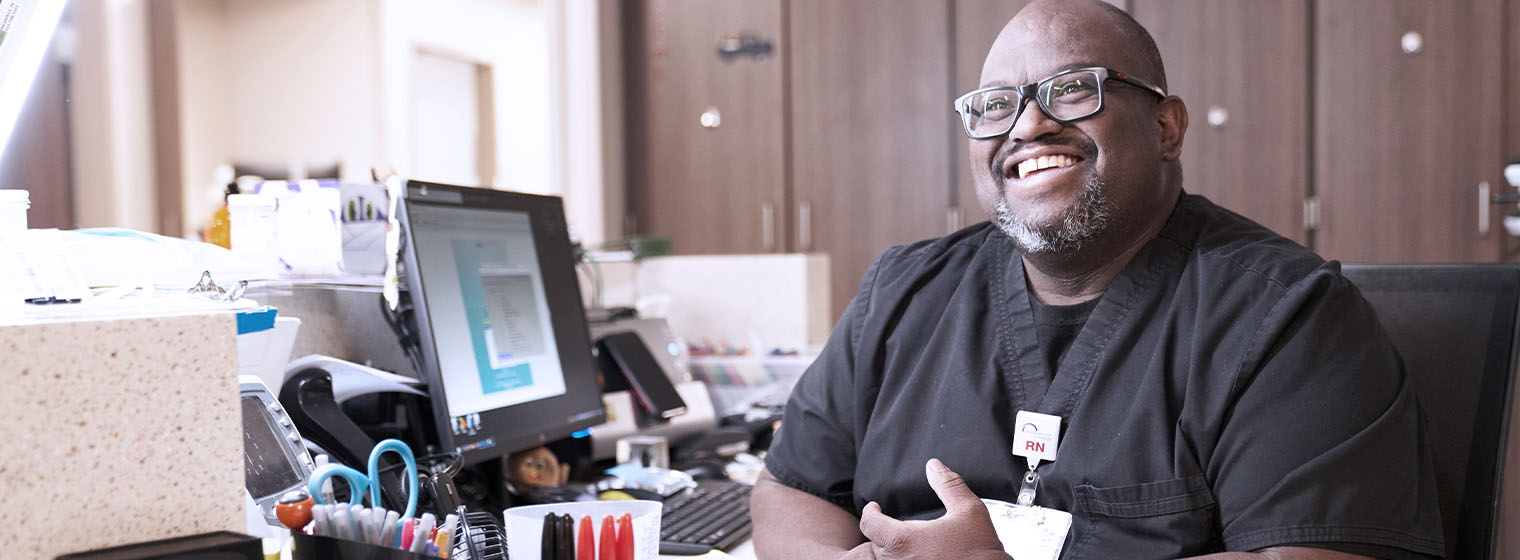 male RN in black scrubs sitting at a computer smiling