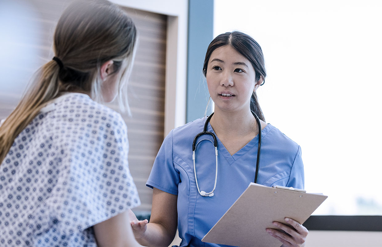 young female nurse with clipboard talking to patient in hospital gown