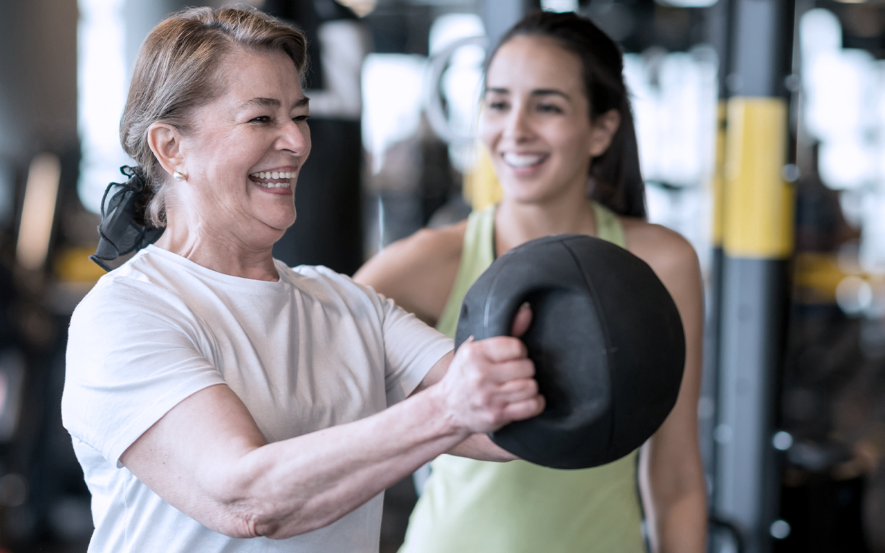young female trainer smiling while elderly female patient lifts medicine ball