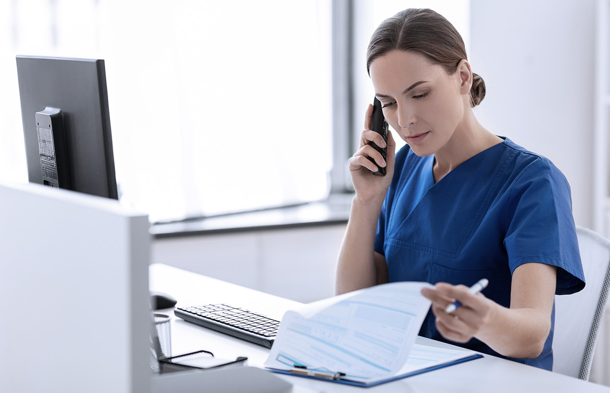 female nurse sitting at computer on the phone looking through paperwork