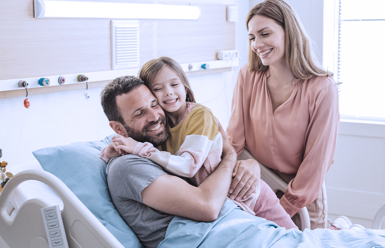 toddler girl hugs her dad in a hospital bed with mom smiling at them