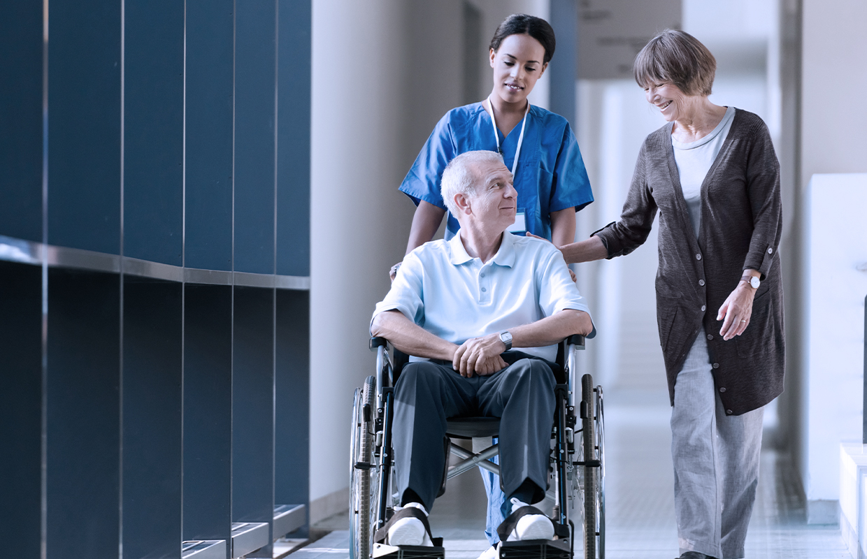 elderly male in a wheelchair being pushed down a hallway with a surgeon and his wife