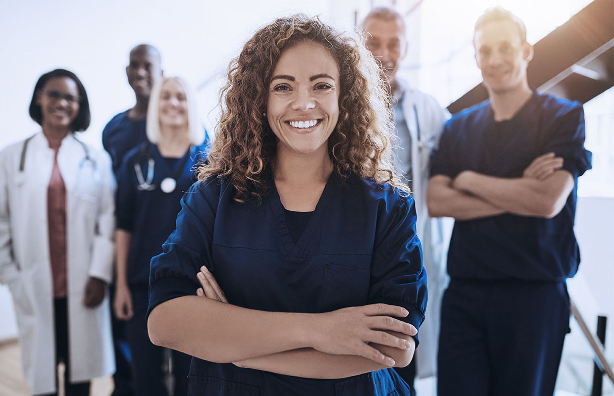 smiling female nurse with arms crossed with more medical staff smiling behind her