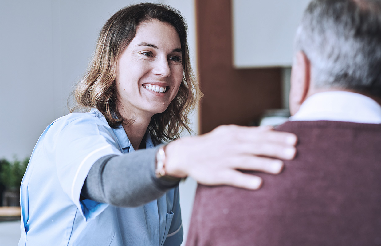 adult female nurse patting the back of an elderly male patient in his home