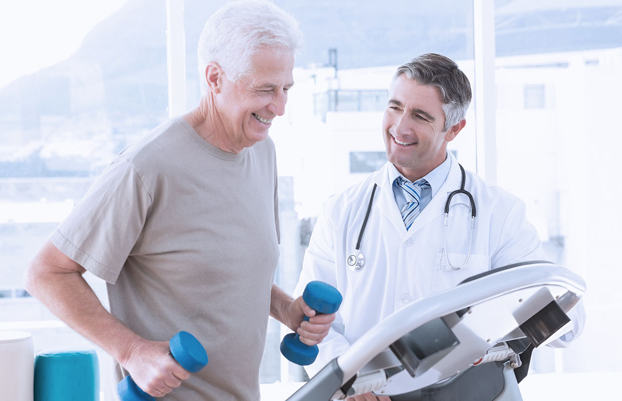 An elder man on elliptical holding weights in both his hands while a doctor smiles at him.