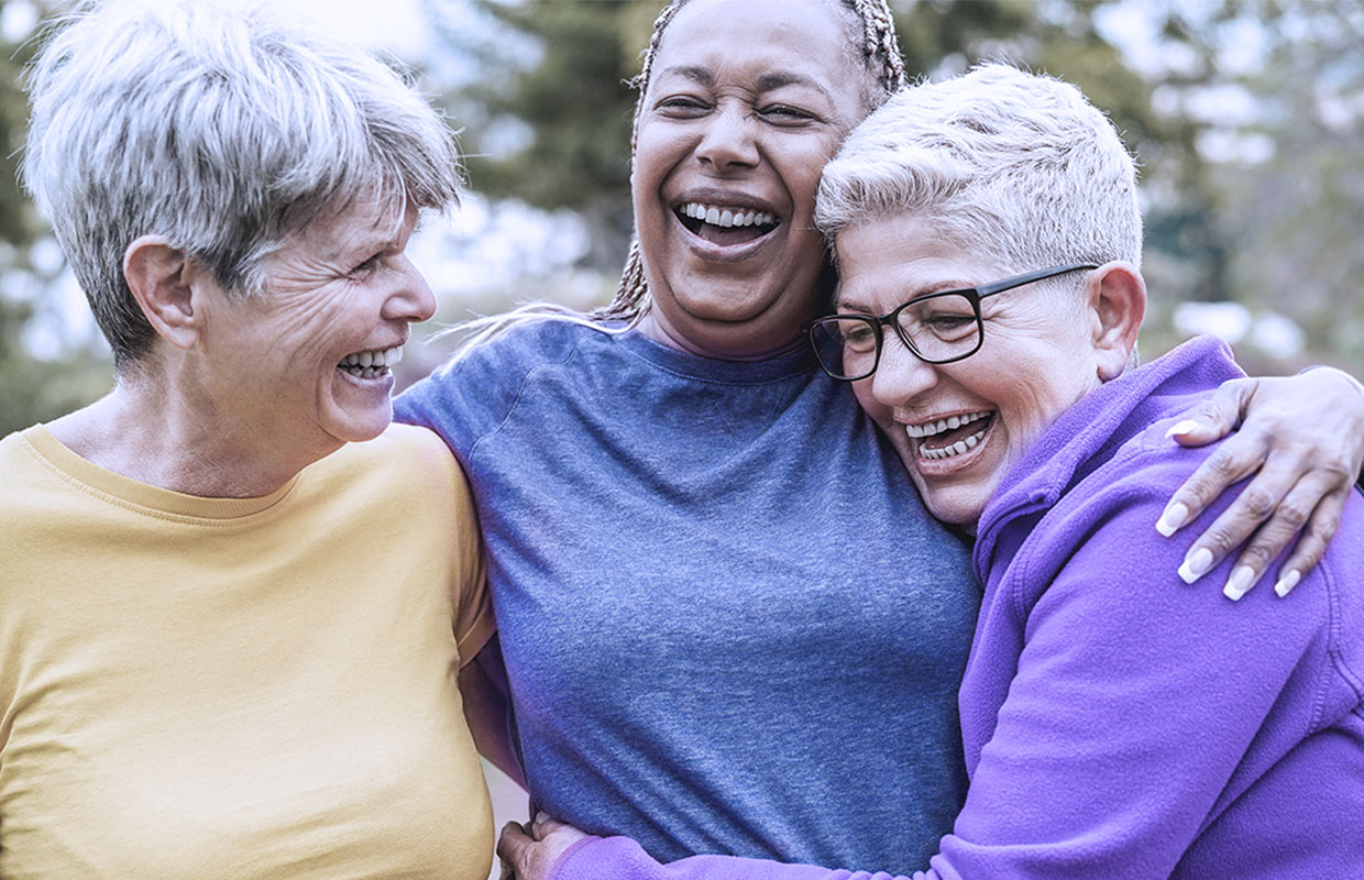 Three female friends in their 60s laugh and hug while walking outsdde.