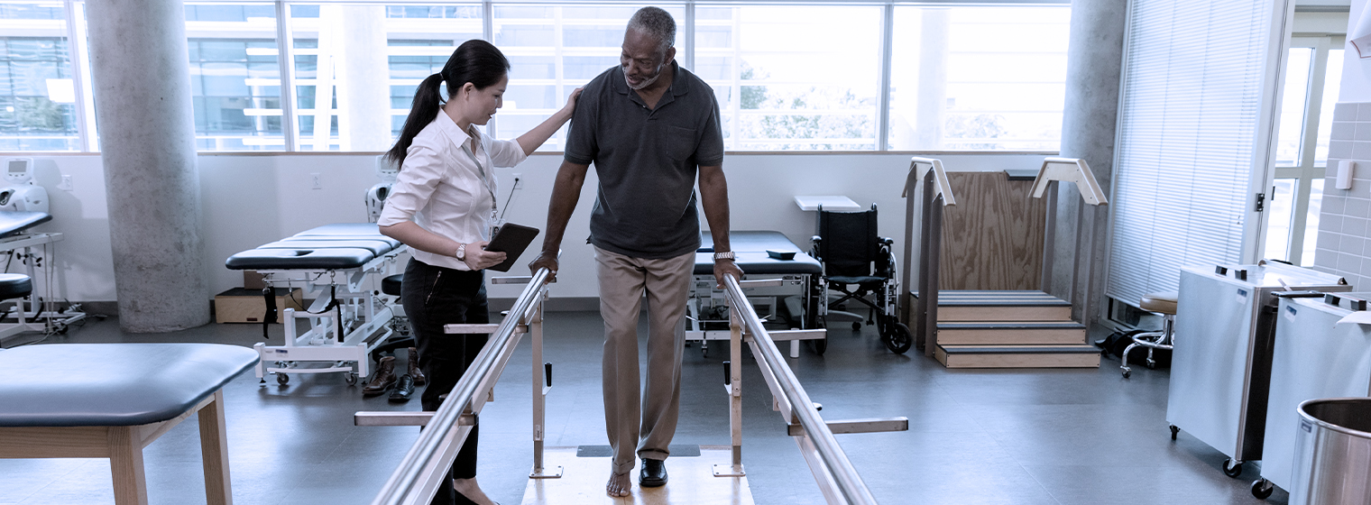 Female provides rehabilitation services to elderly male using parallel bars.