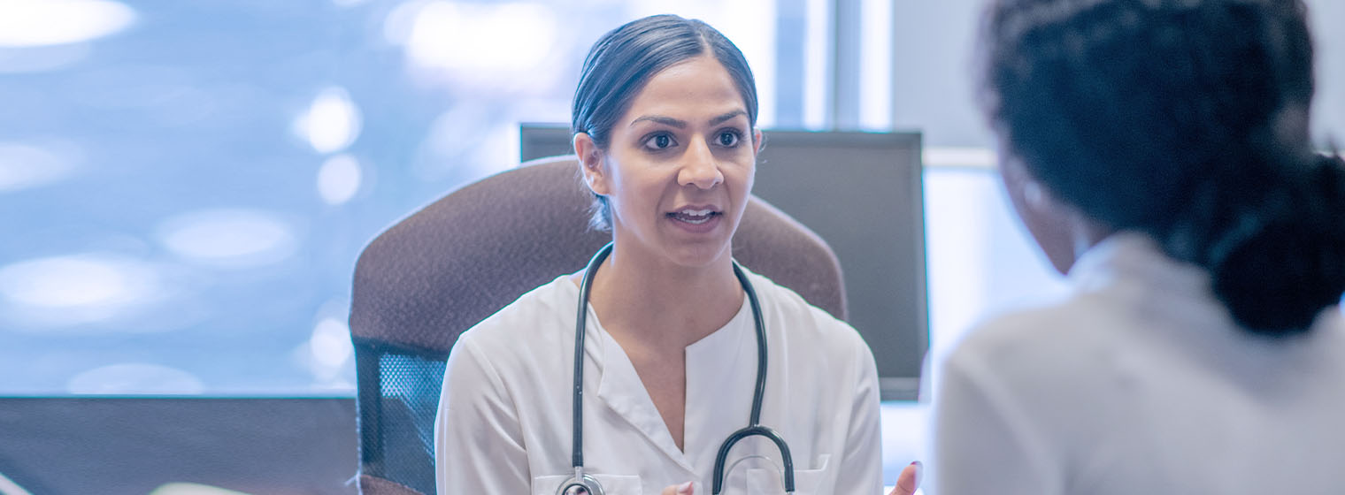 Female doctor sits on a stool while she talks to a female patient regarding her healthcare needs.