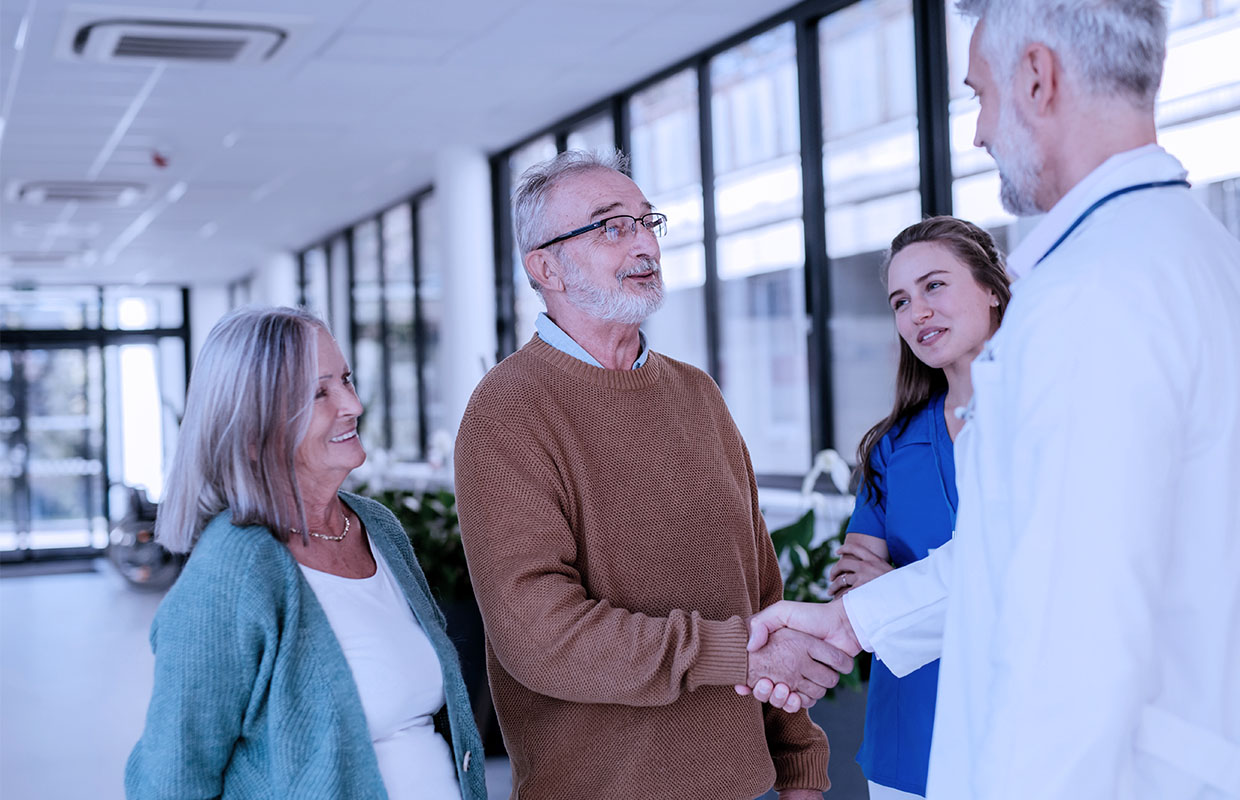 elderly male shakes elderly doctor's hand while standing next to his wife and young female nurse