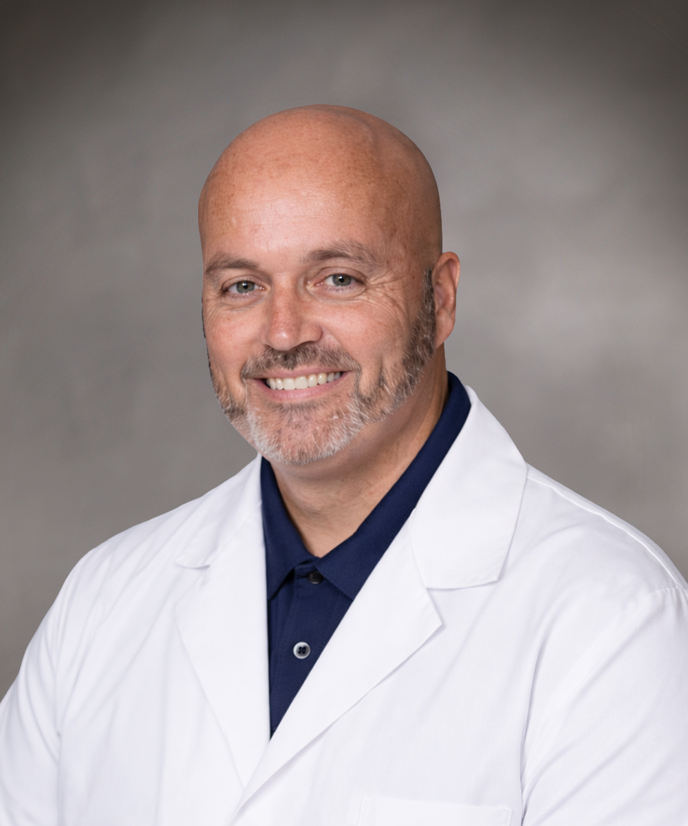 A smiling, middle-aged bald man with a short beard is wearing a white lab coat over a dark collared shirt, representing labor and delivery in Morristown, and poses against a neutral gray background.