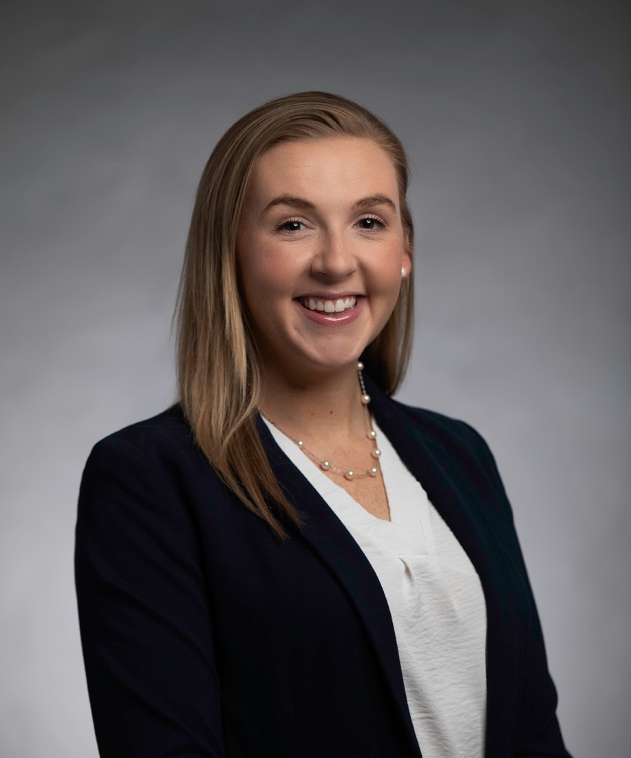 A young woman with straight, light brown hair wearing a dark blazer over a white blouse and a delicate necklace, smiling at the camera against a neutral gray background—an ideal fit for nursing jobs at Parkwest Medical Center.
