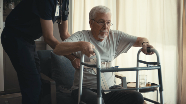 An older man using a walker is assisted by a caregiver as he gets up from a chair, illustrating compassionate hospice care in an indoor setting with soft lighting and a glass of water on a nearby table.
