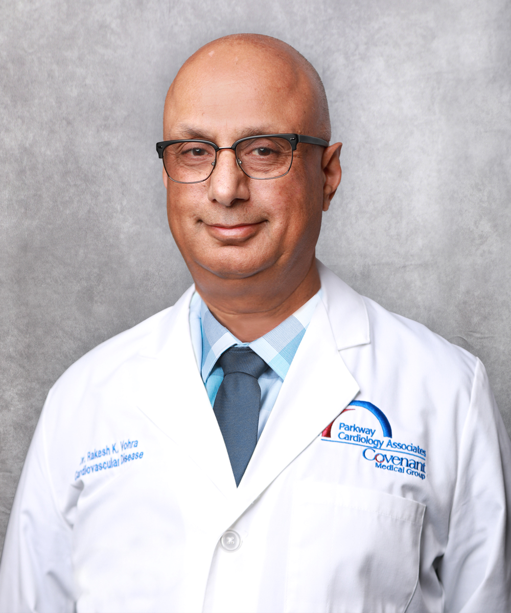 A bald man wearing glasses, a white doctor’s coat with the Covenant Medical Group logo, and a name badge smiles softly against a gray background, reflecting his expertise in cardiology. He wears a blue plaid shirt and dark tie.