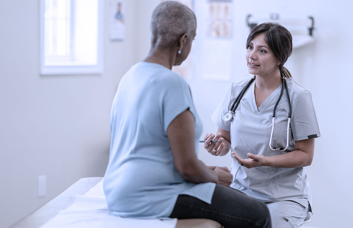 middle age female patient sits on bed in doctor's office talking to young female nurse