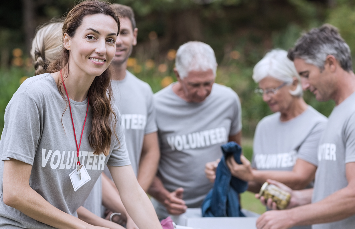 a woman smiles for a picture while a group of volunteers behind her collect food and clothes for donation